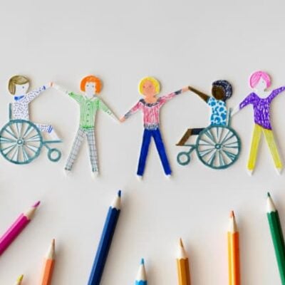 supporto bianco nella cui parte inferiore sono presenti delle matite colorate. Al di sopra di esse cinque persone che giocano e si divertono. Due di queste sono in sedia a rotelle. Lo stile dei personaggi è disegnato a mano e ritagliato, sono delle forme di carta