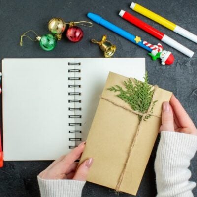 quaderno bianco con pennarelli e matite attorno. In primo piano una busta con un pezzetto di pino attaccato