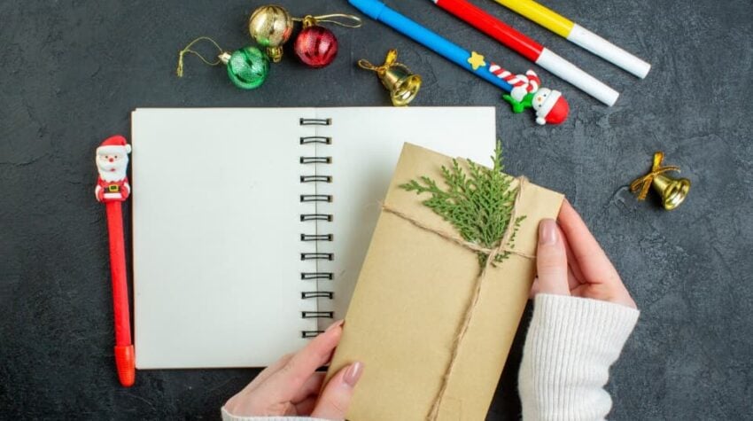 quaderno bianco con pennarelli e matite attorno. In primo piano una busta con un pezzetto di pino attaccato